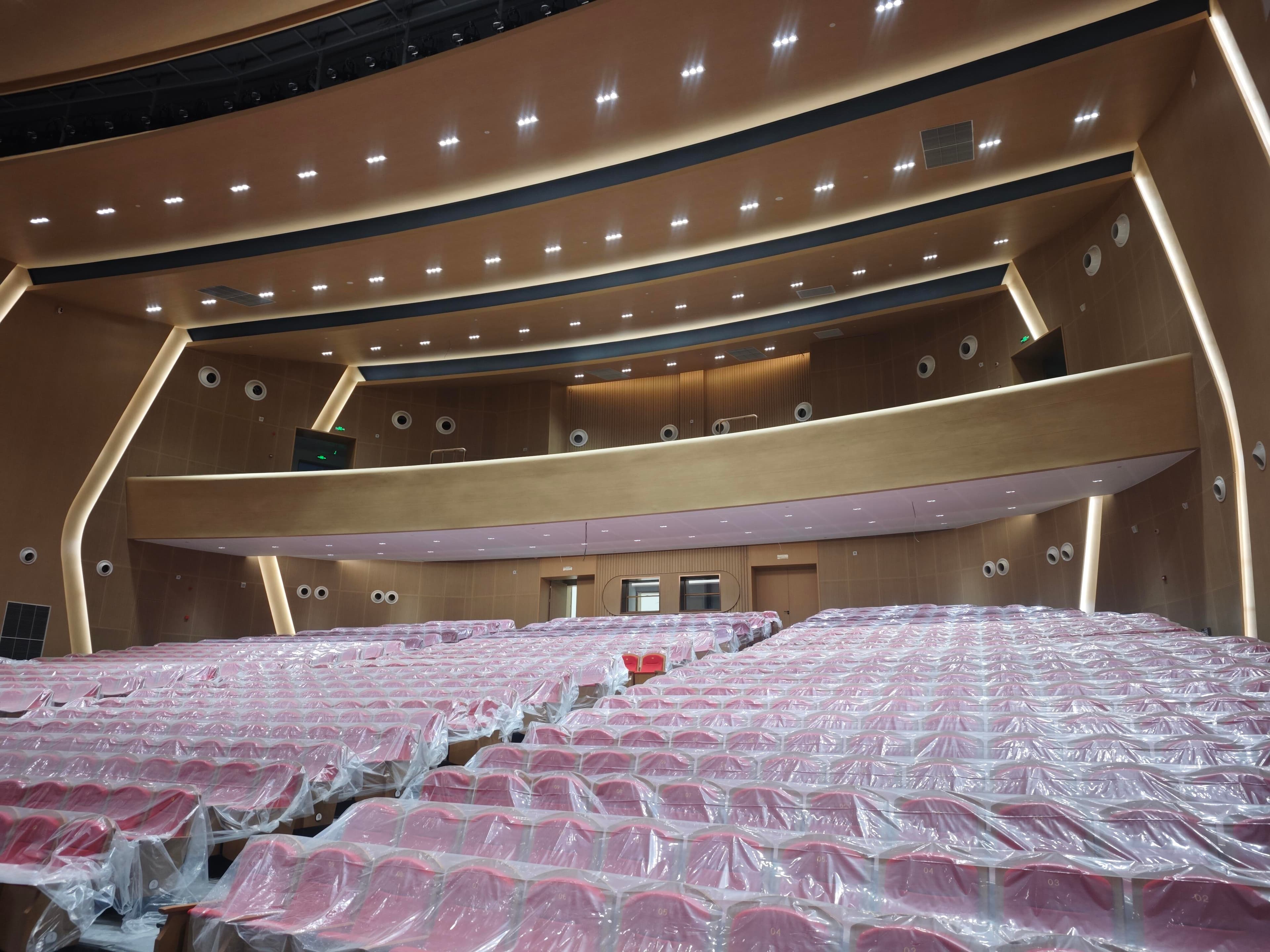 Auditorium seating area with red fabric seats, balcony, and warm wood-tone acoustic finishes