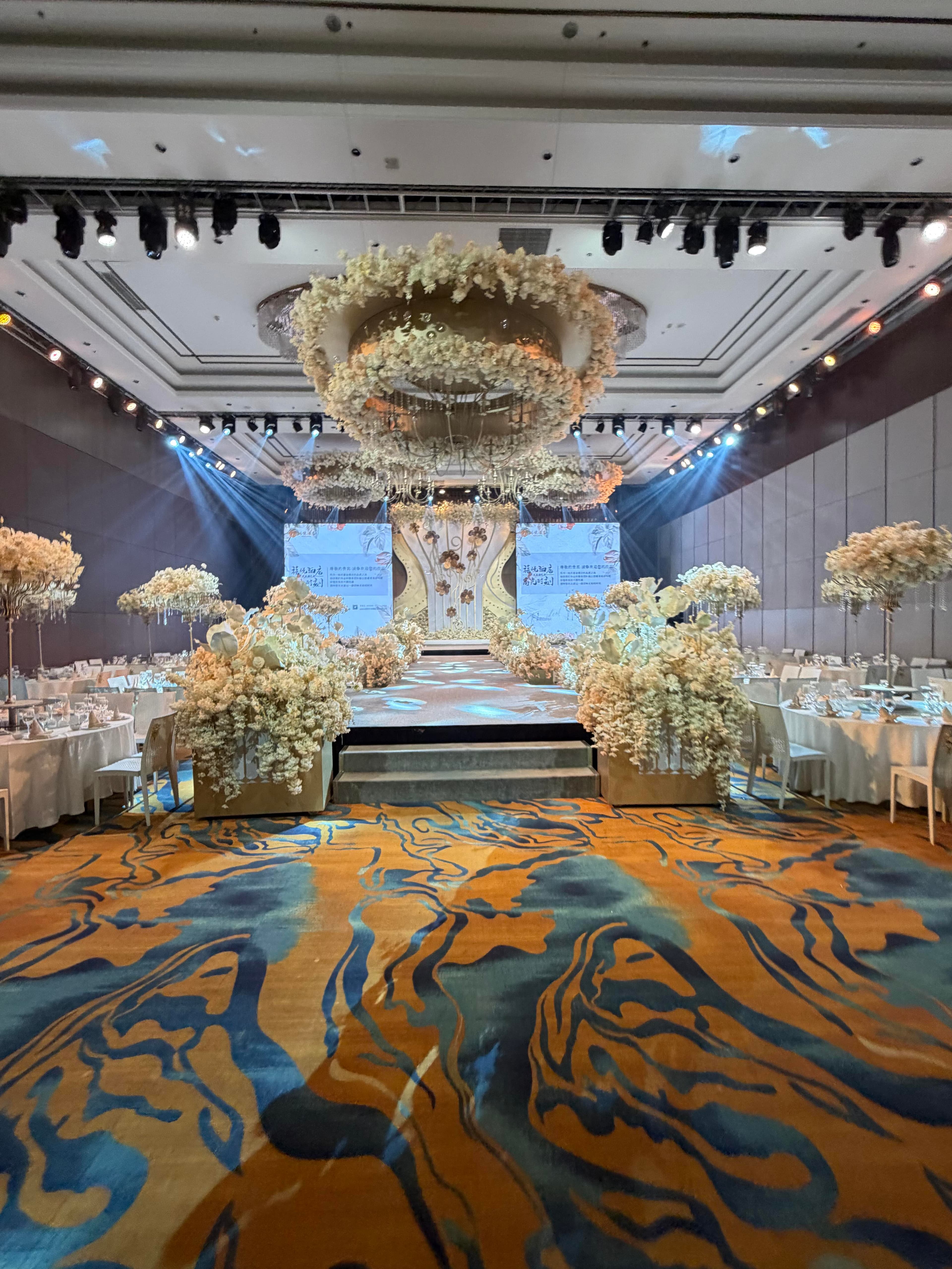 Golden warm-toned ballroom with low-height floral wreath and stage visual center