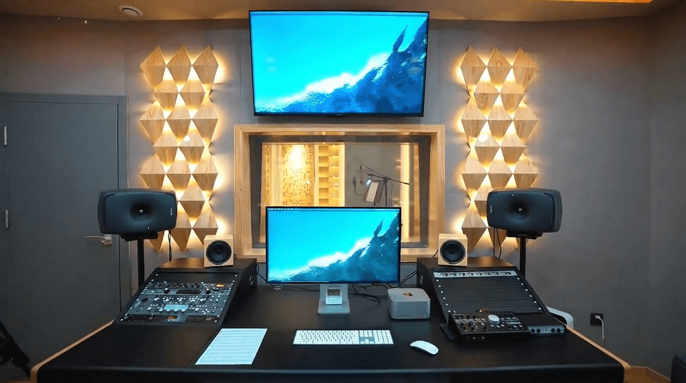 Control room front workspace with custom black desk, dual displays, outboard gear racks, monitors, and observation window